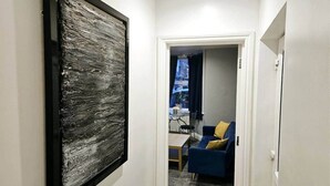 Un pasillo con un cuadro abstracto grande a la izquierda, un sofá azul con un cojín amarillo al fondo, y una puerta que da a otra habitación.