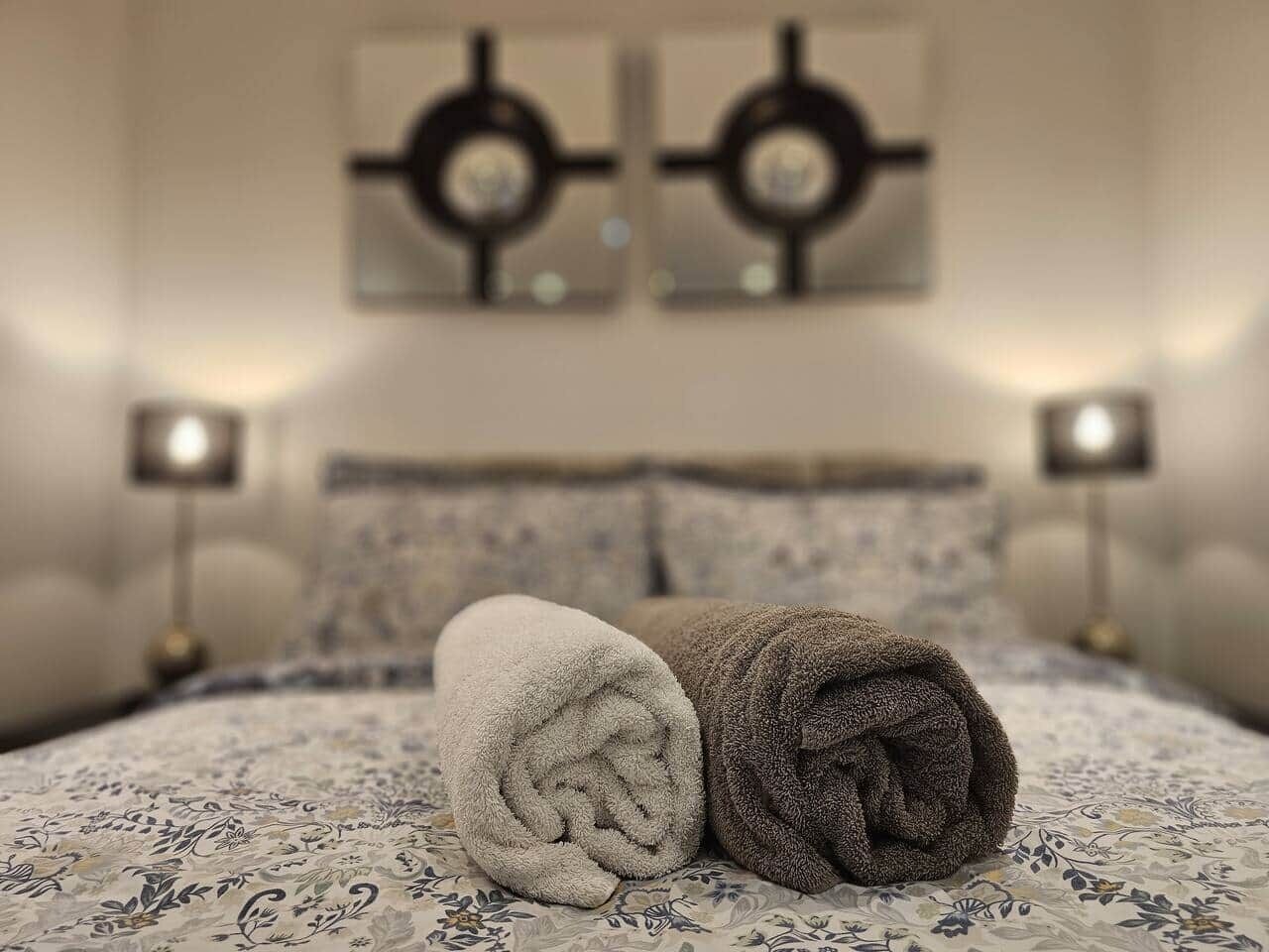A neatly made bed with rolled towels, a patterned bedspread, and two wall-mounted lamps.
