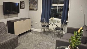 Una sala de estar moderna con un televisor de pantalla plana, un sofá, una mesa de centro de vidrio con una planta y un comedor con mesa y sillas.