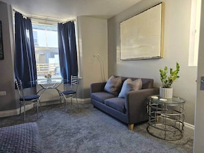 A living room with a grey sofa, a glass coffee table, a small round table with a plant, and a view of the outside through large windows with blue curtains. - Stay Zen Apartments - Free Parking & Garden Woking (Woking)