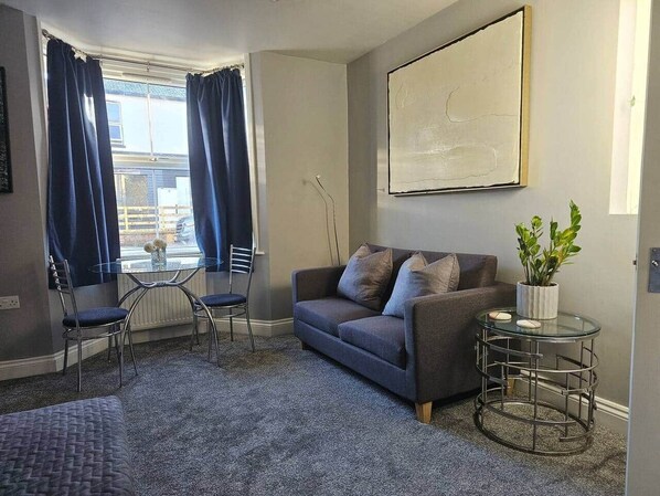 A living room with a grey sofa, a glass coffee table, a small round table with a plant, and a view of the outside through large windows with blue curtains.