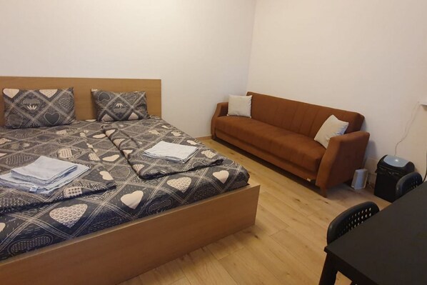 A room with a bed, a brown sofa, a small table, and a trash bin.