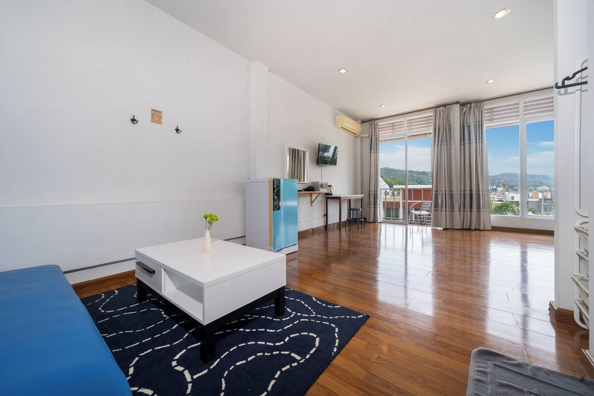 A modern hotel room with a wooden floor, a white desk, a blue sofa, a small table with a vase, a TV on the wall, and a large window with a view of the city.