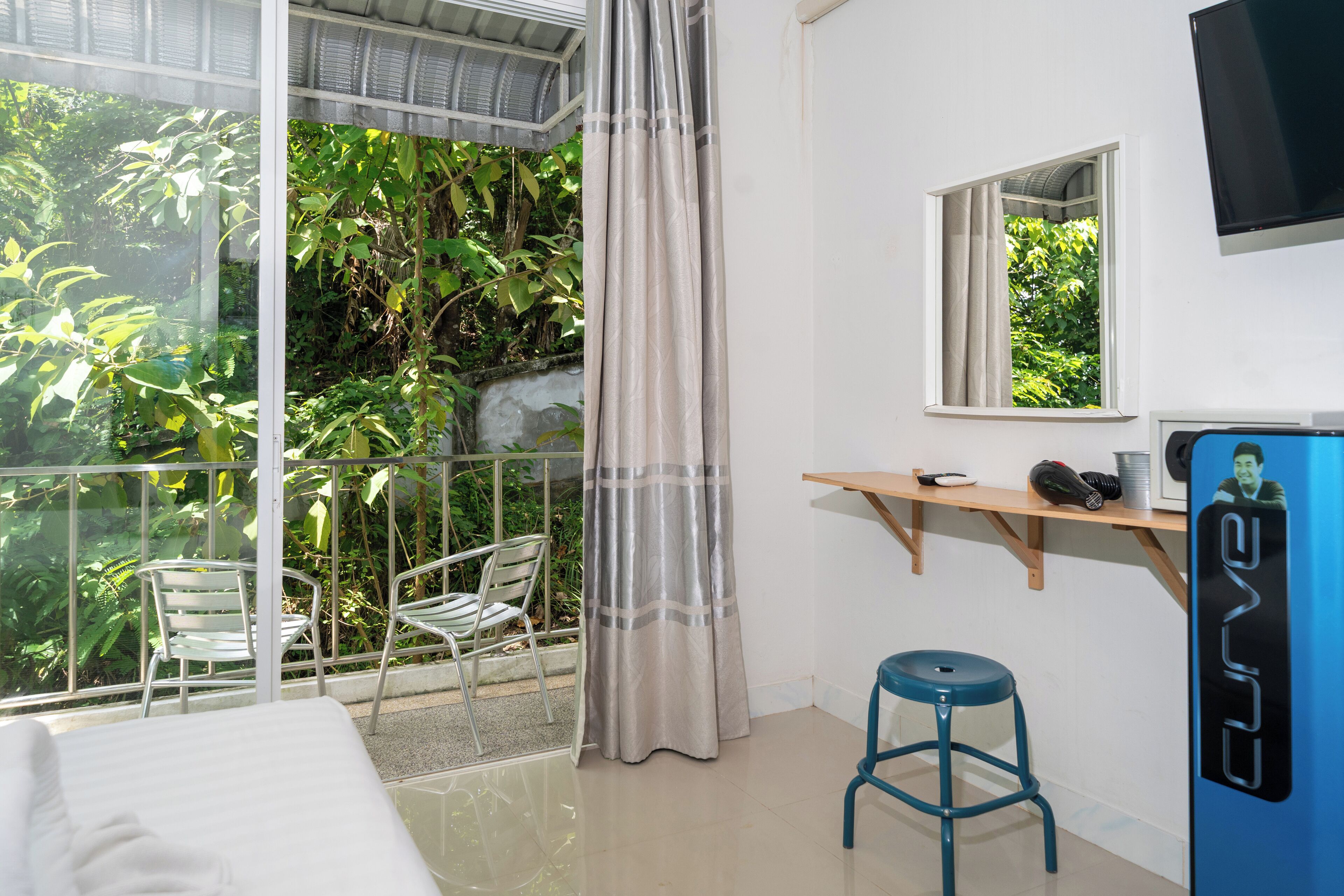 A room with a bed, a desk with a chair, a TV, and a view of greenery through large windows.
