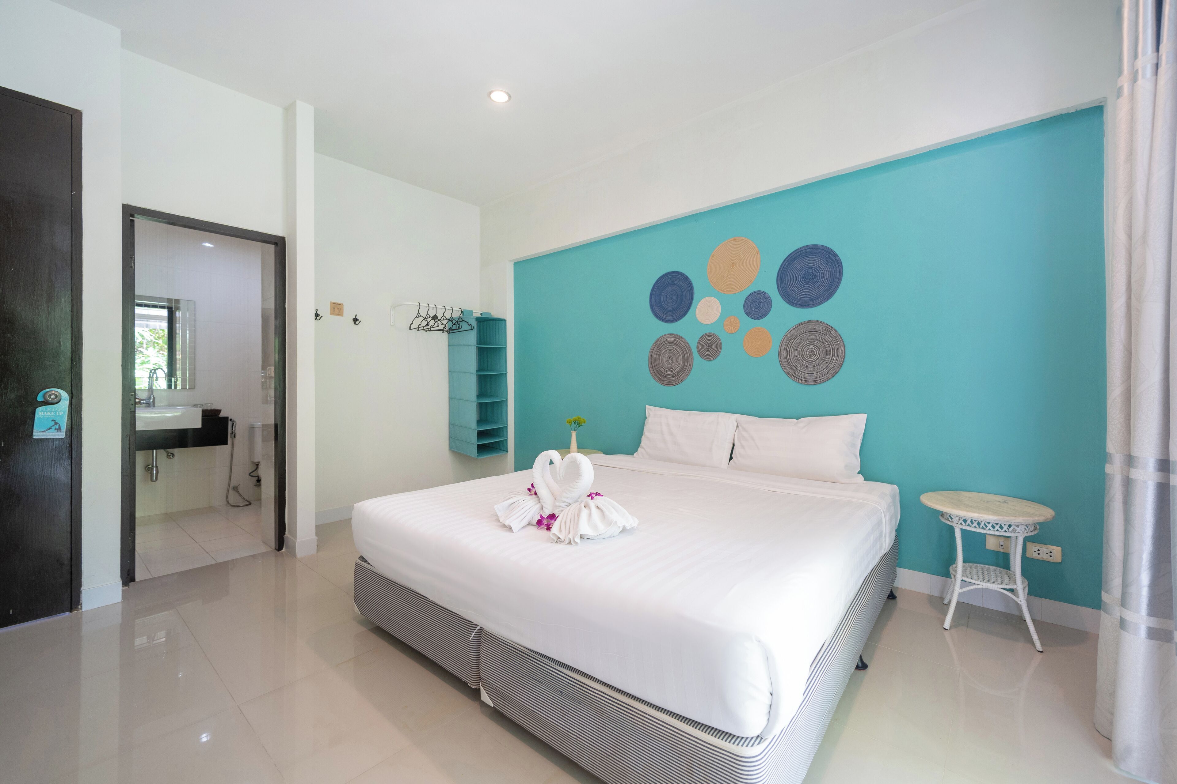 A modern hotel room with a large bed, a small round table, a bathroom visible through an open door, and a teal accent wall.