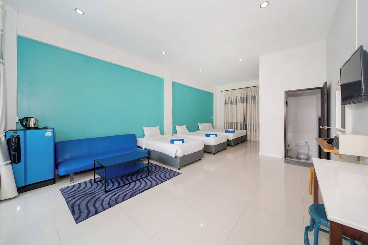 A hotel room with three beds, a blue sofa, a small kitchen area, and a TV.