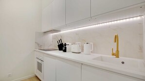 A modern kitchen with white cabinetry, a marble backsplash, a built-in oven, a coffee maker, a toaster, and a gold faucet. - Charming 3-bedroom Flat - Le Marais (Paris)