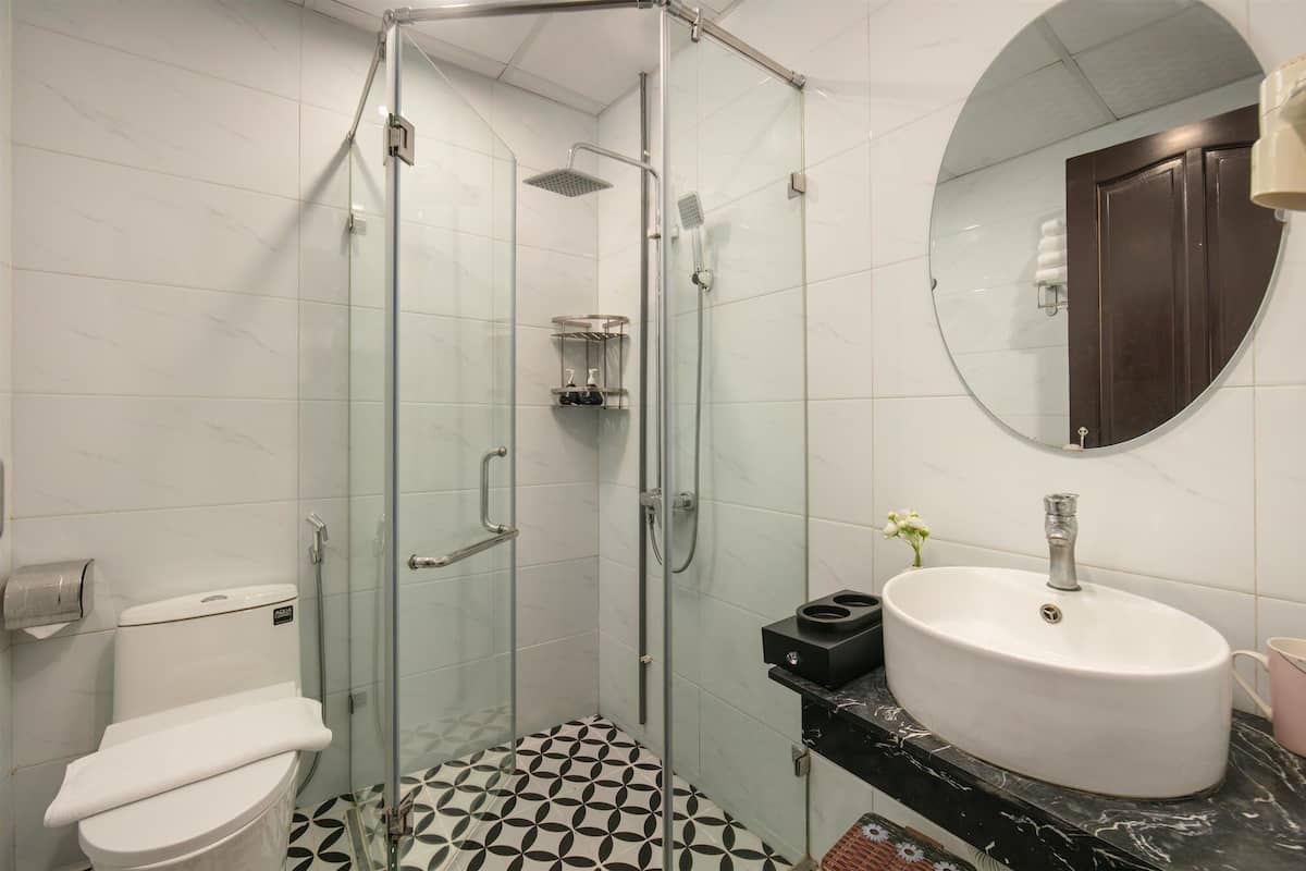 A bathroom with a glass-enclosed shower, a toilet, a sink with a marble countertop, and a round mirror.