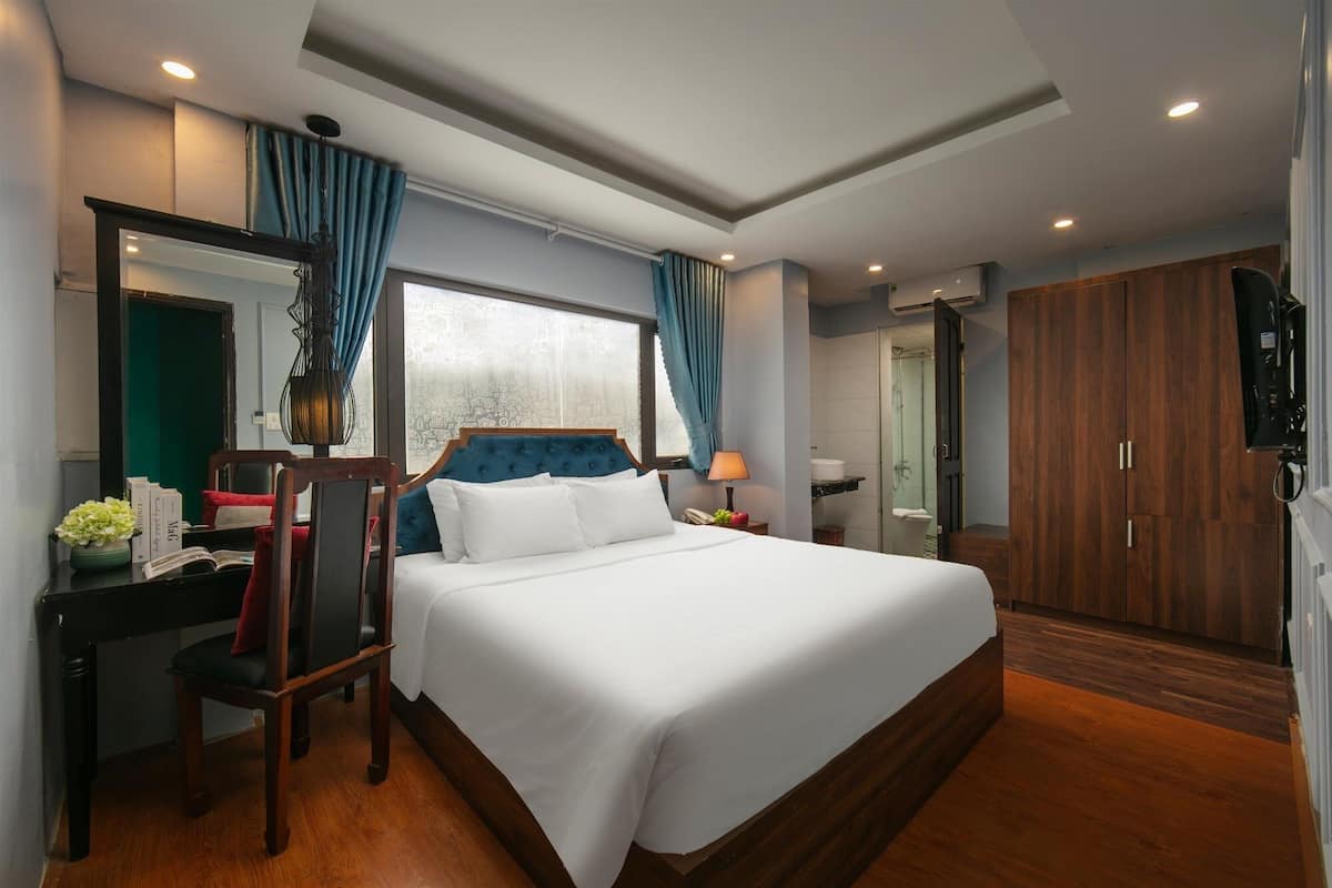 A hotel room with a large bed, a desk with a chair, a wardrobe, and a bathroom visible through an open door.