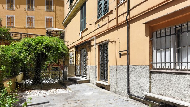 Un étroit sentier de pierres mène à un bâtiment à la façade jaune, doté d’un portail en métal et d’un petit banc.
