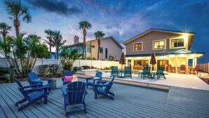 Una terraza con asientos al aire libre y zona de piscina, rodeada de palmeras y una casa con porche cubierto.