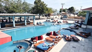 Zona de piscina con sillones de descanso y pufs, rodeada de árboles y un edificio.