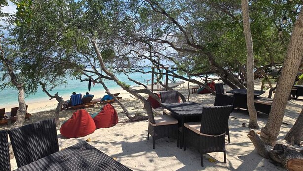 Un área de estar frente a la playa con sillas y mesas de mimbre, un bote pequeño y un letrero al fondo.