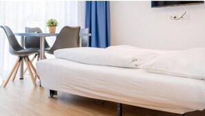 Un lit simple avec un matelas blanc et un cadre en métal noir, une petite table avec des chaises, une plante en pot et une fenêtre avec des rideaux bleus.