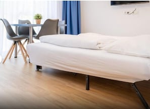 A single bed with a white mattress and black metal frame, a small table with chairs, a potted plant, and a window with blue curtains.
