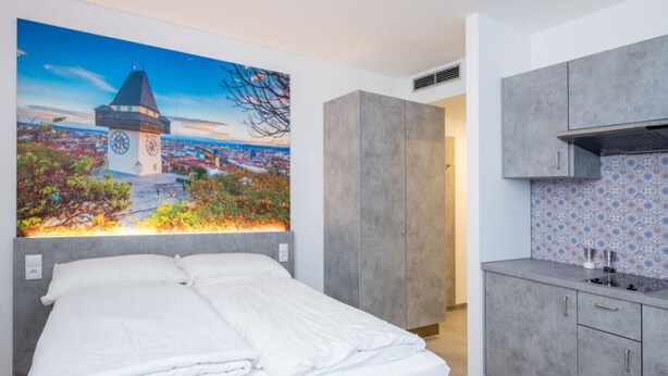 Une chambre d’hôtel moderne avec un grand lit, une petite kitchenette et une fresque représentant un paysage urbain sur le mur.