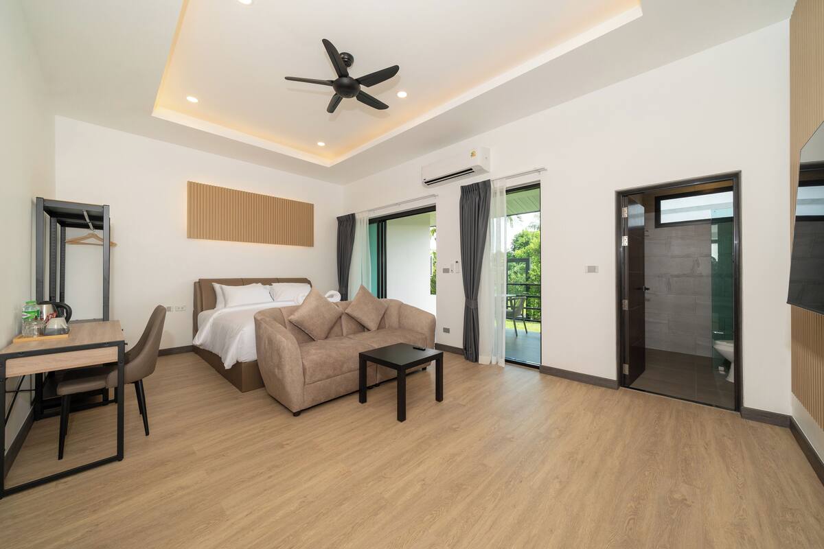 A modern hotel room with a bed, sofa, dining table, and chairs. There is a ceiling fan, an air conditioning unit, and a view of the outdoors through a glass door.