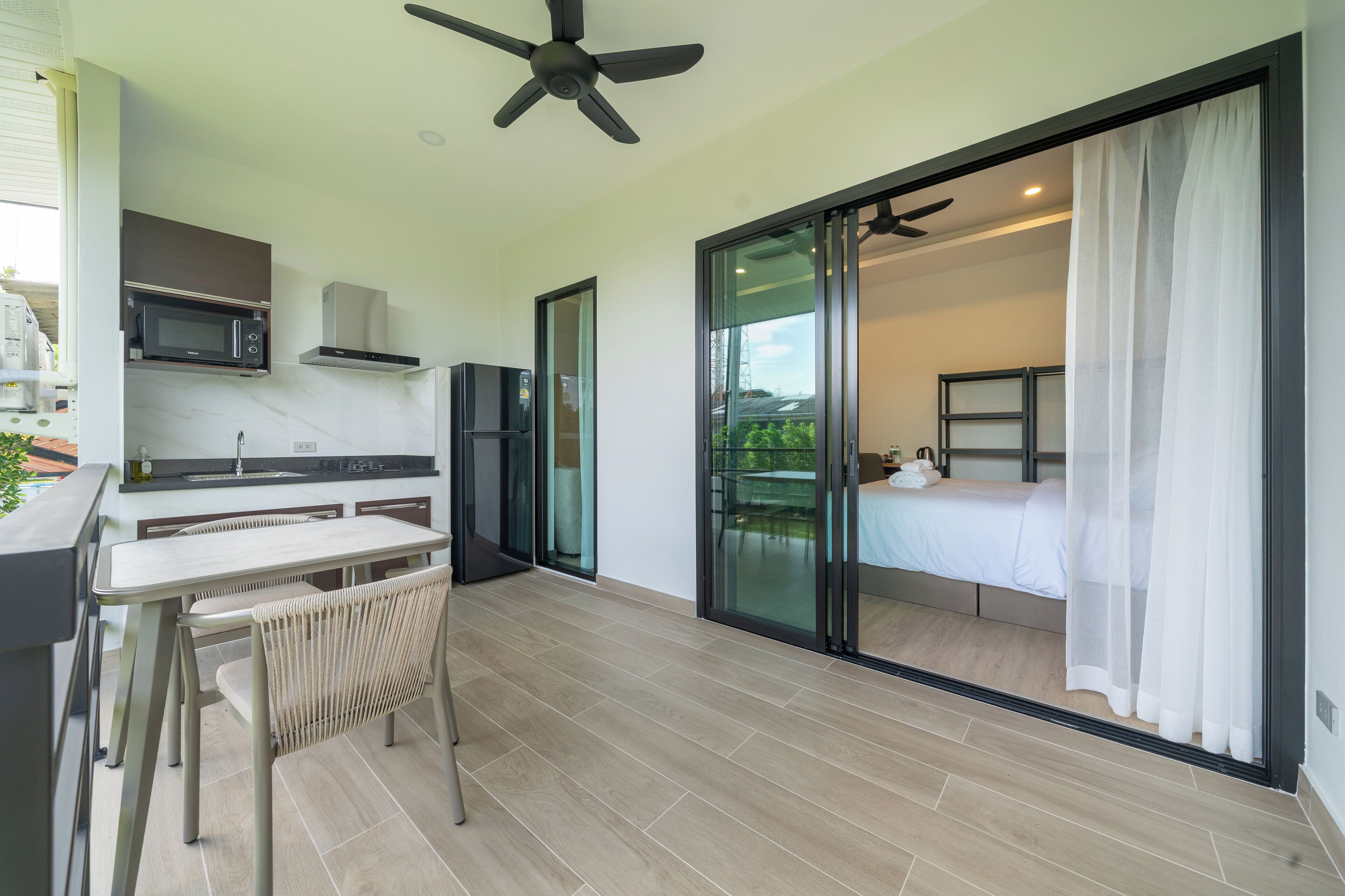 A modern kitchen with a microwave, stove, and water dispenser. There is a dining area with a table and chairs. The room has a sliding glass door leading to a balcony with a bed.