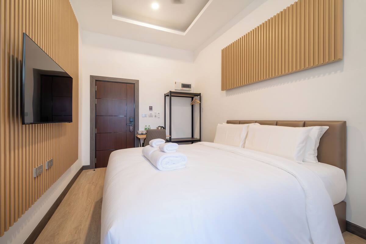 A hotel room with a large bed, a flat-screen TV mounted on the wall, a wooden panel wall, and a door leading to the bathroom.