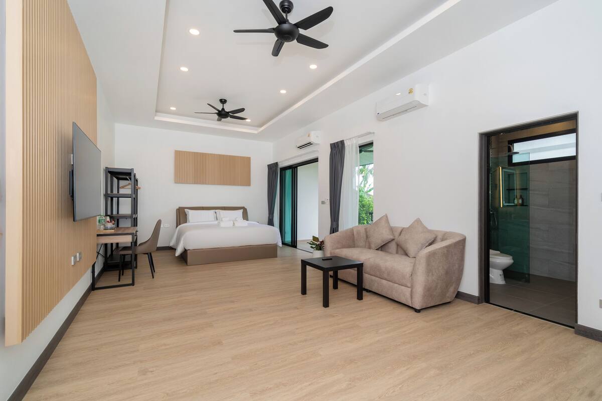 A modern hotel room with a bed, a sofa, a desk, and a TV. There are ceiling fans and an air conditioning unit.