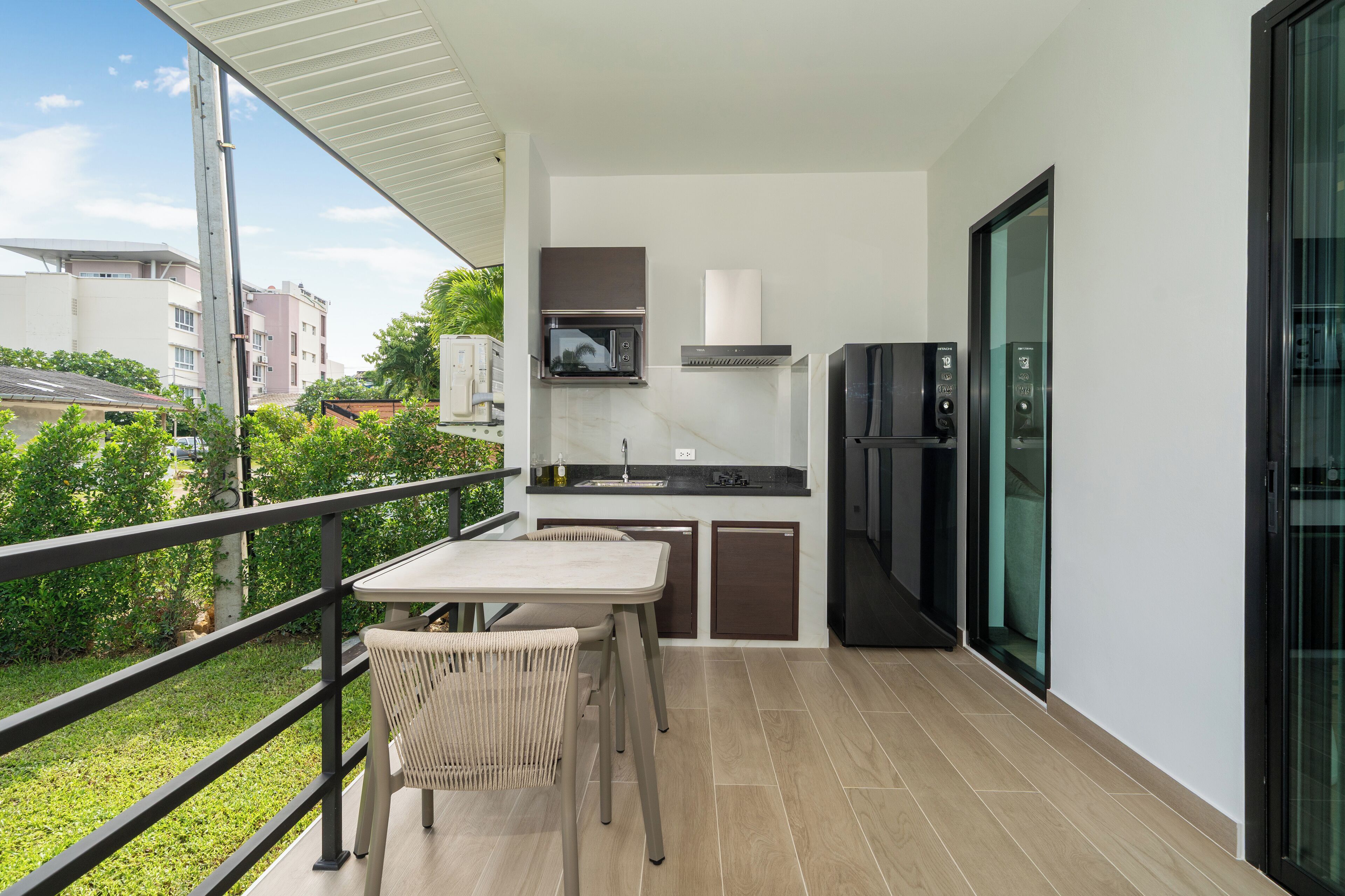 A balcony with a table and chairs, a kitchen area with a microwave, stove, and refrigerator, and a view of greenery and buildings.