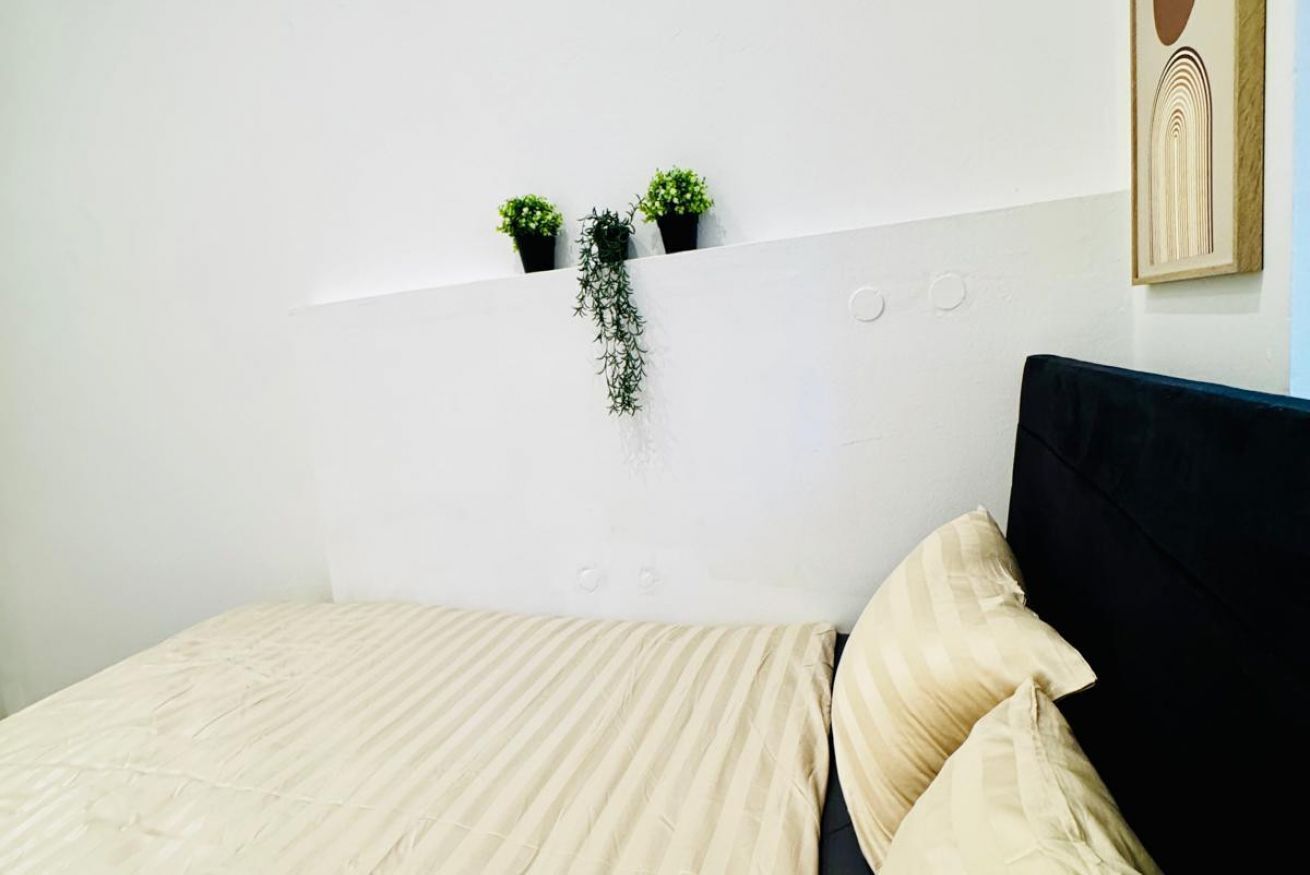 Una cama con cabecera negra, colcha color beige y dos almohadas color beige. Encima de la cama, en la pared, hay tres macetas pequeñas con plantas.
