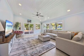 A spacious living room with a large beige sectional sofa, a wooden coffee table, and a TV on a wooden stand. There are large windows allowing natural light and views of the outdoors.