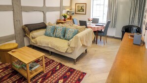 A cozy room with a bed, a small table, a desk, and a chair. There is a window with curtains and a painting on the wall.