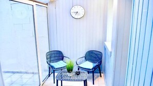 Een balkon met twee rieten stoelen, een bijzettafeltje en een wandklok.