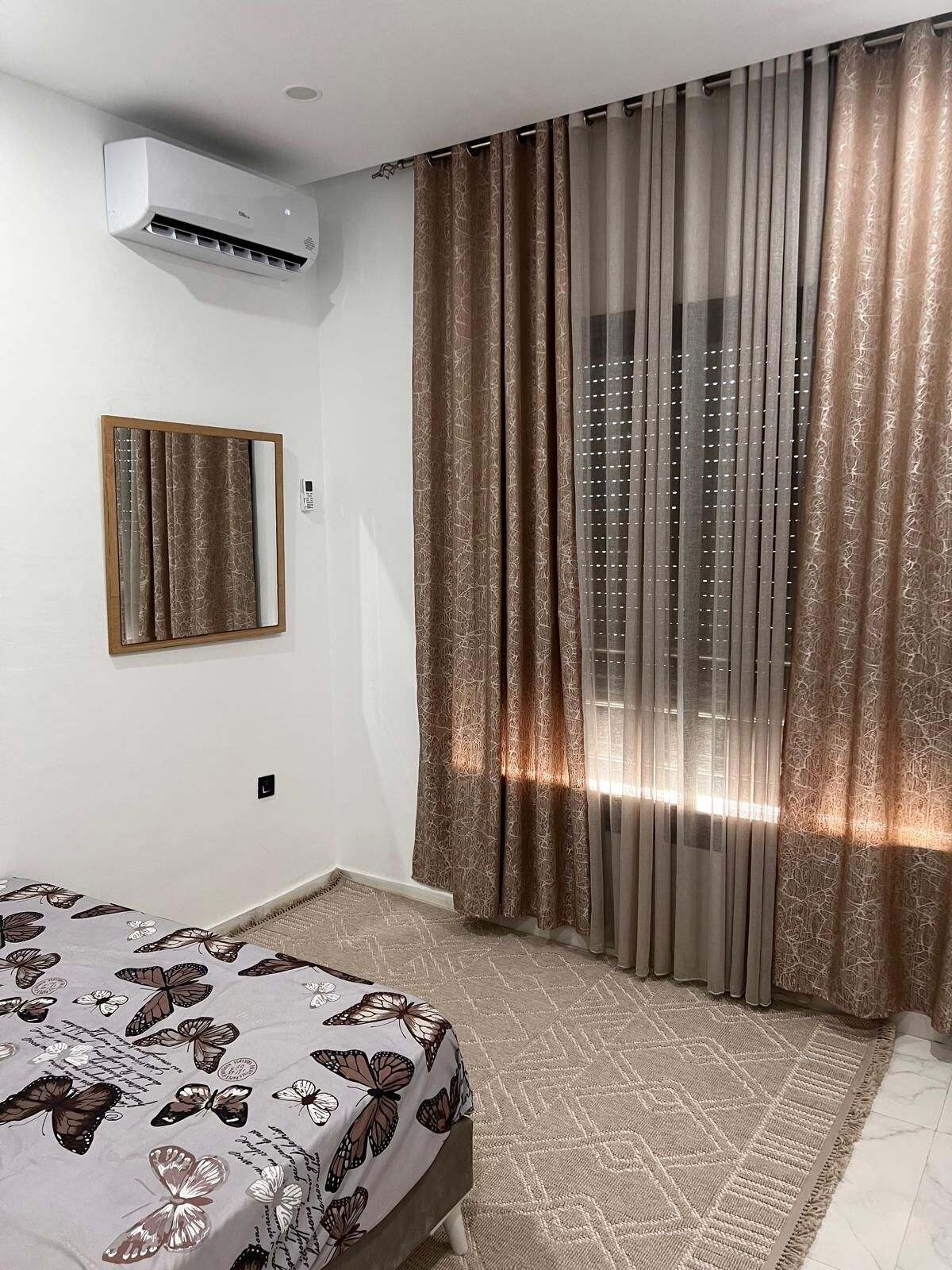 A bedroom with a bed, a window with curtains, an air conditioning unit, and a mirror on the wall.