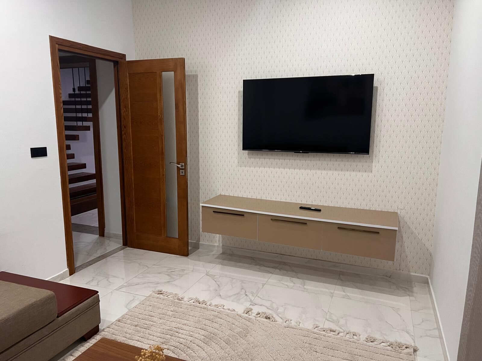 A room with a TV mounted on the wall, a wooden door, a marble floor, and a beige console table.