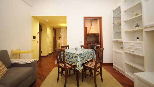 Un comedor con una mesa dispuesta para cuatro personas, rodeada de sillas de madera. Un sofá se encuentra a la izquierda y una estantería blanca a la derecha.