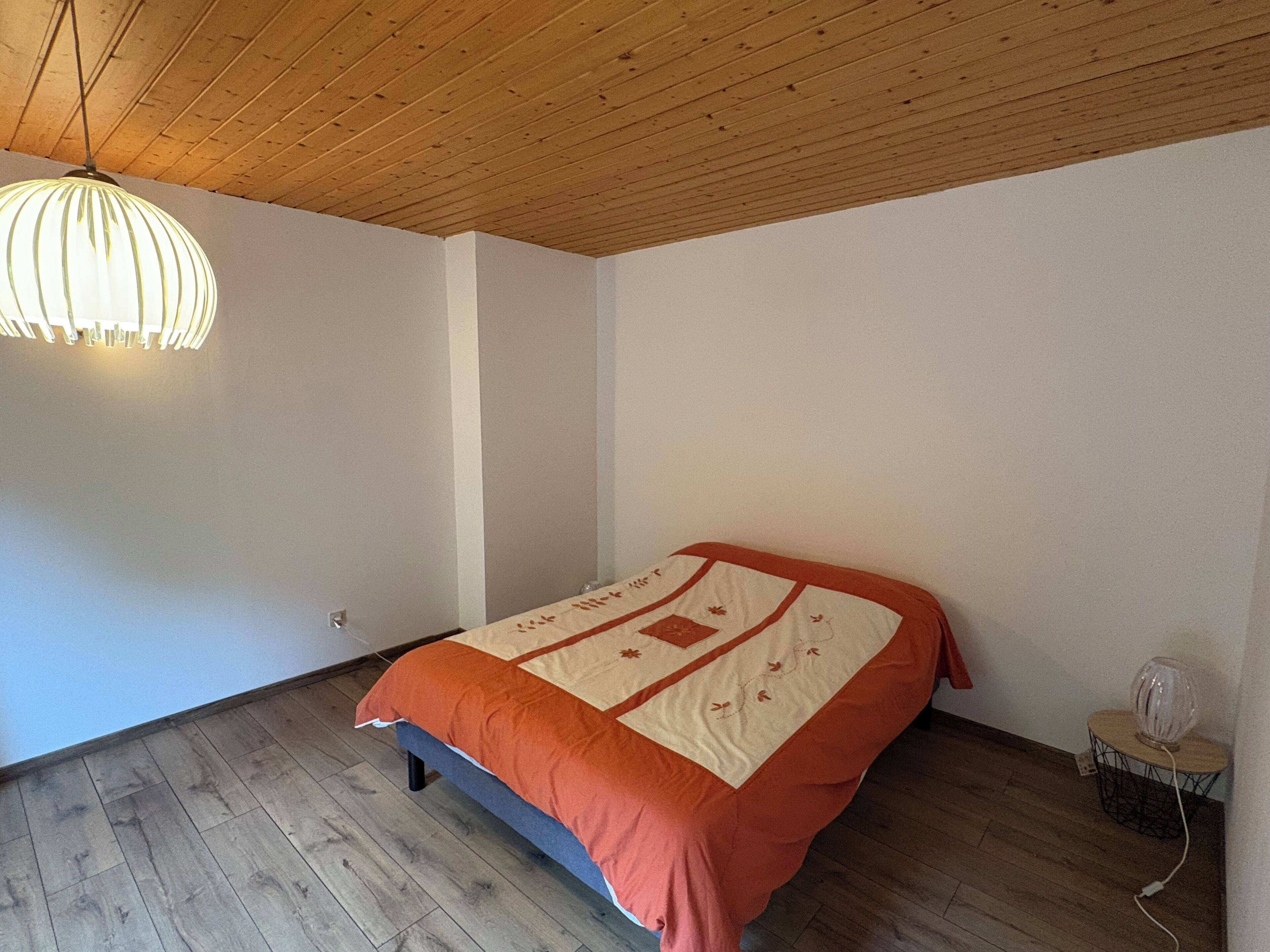A bedroom with a wooden ceiling, a bed with an orange and white bedspread, a hanging light fixture, and a small table with a lamp.