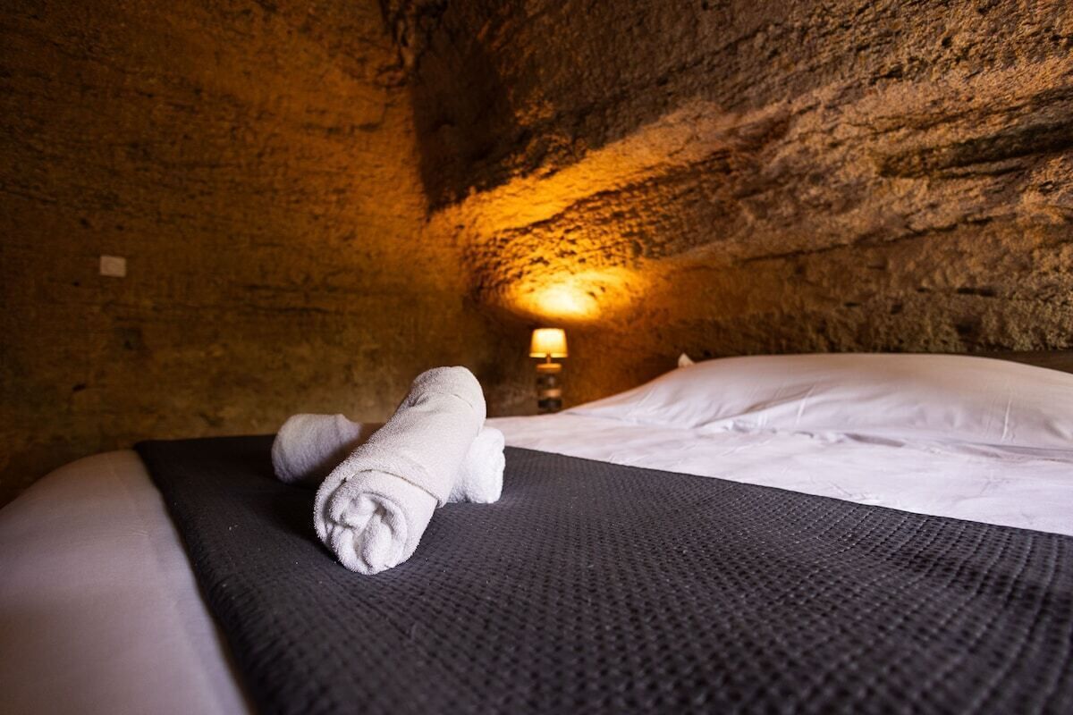 A bed with a rolled towel, a textured blanket, and a lamp in a cave-like room with rough walls.