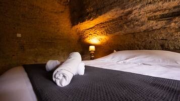 A bed with a rolled towel, a textured blanket, and a lamp in a cave-like room with rough walls.