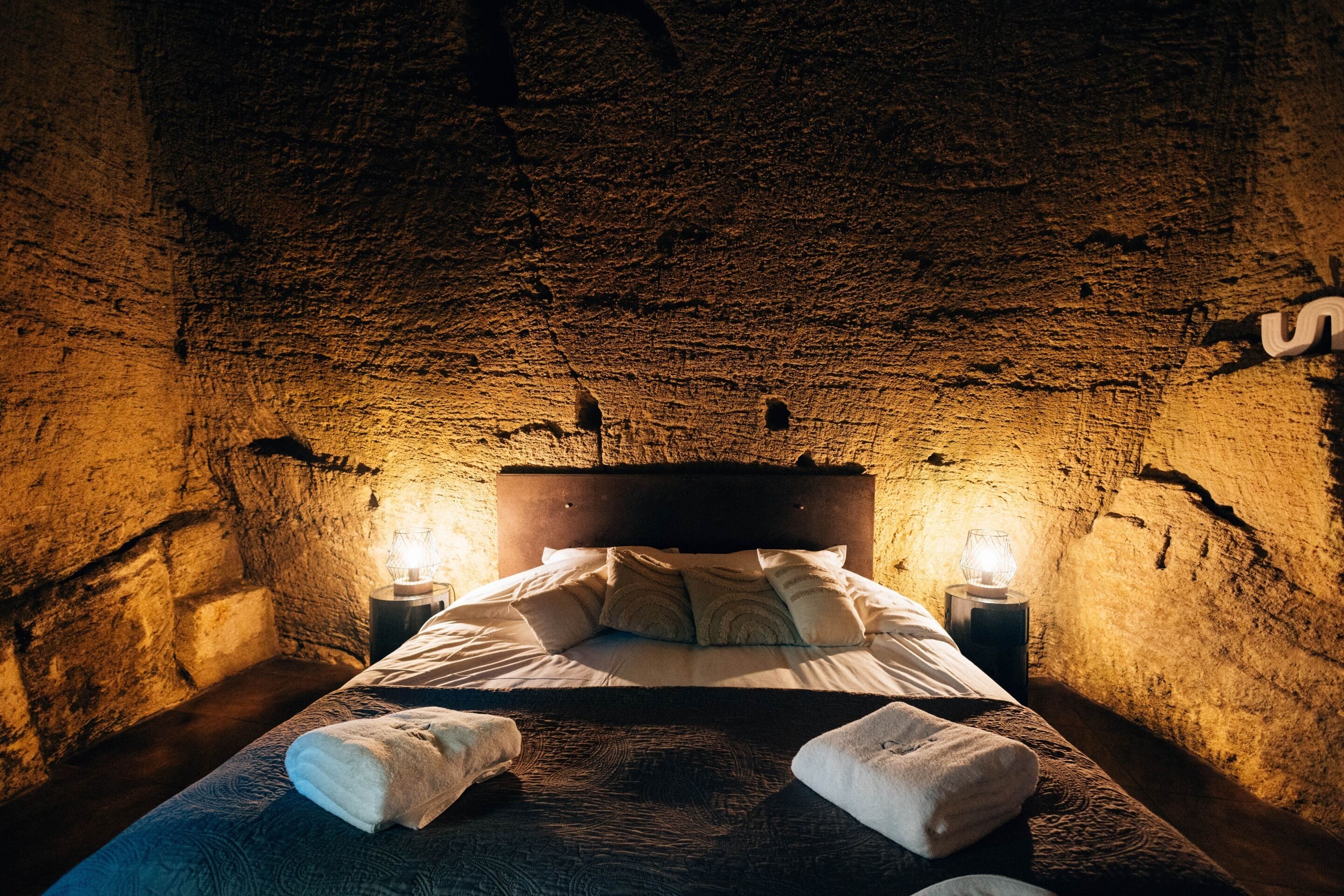 A cozy room with a rustic stone wall, a wooden bed with pillows, and two bedside lamps.