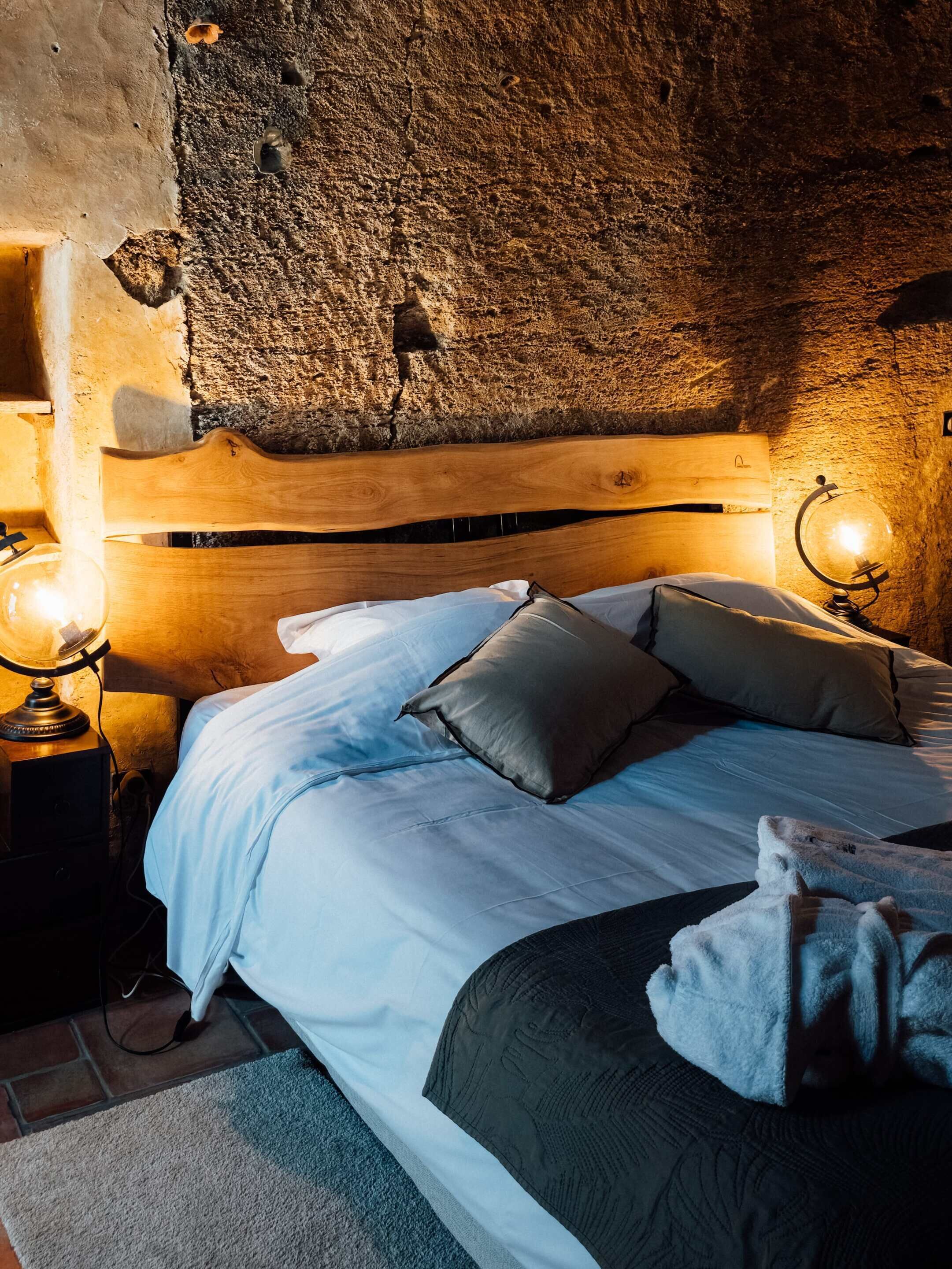 Une chambre confortable avec un mur en pierre rustique, une tête de lit en bois et un lit recouvert de draps blancs et agrémenté de plusieurs oreillers.