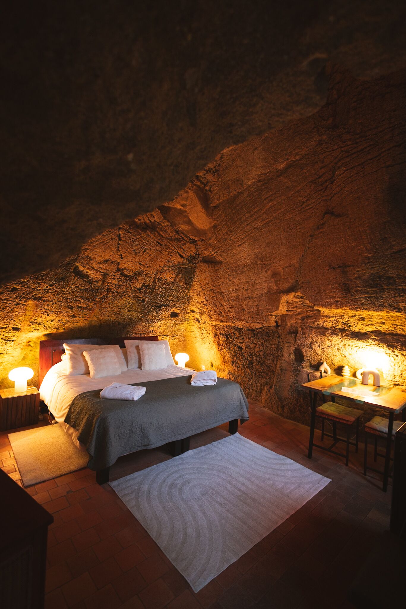 A bedroom with a bed, bedside tables, and a small table with chairs, set in a cave-like room with natural rock walls.