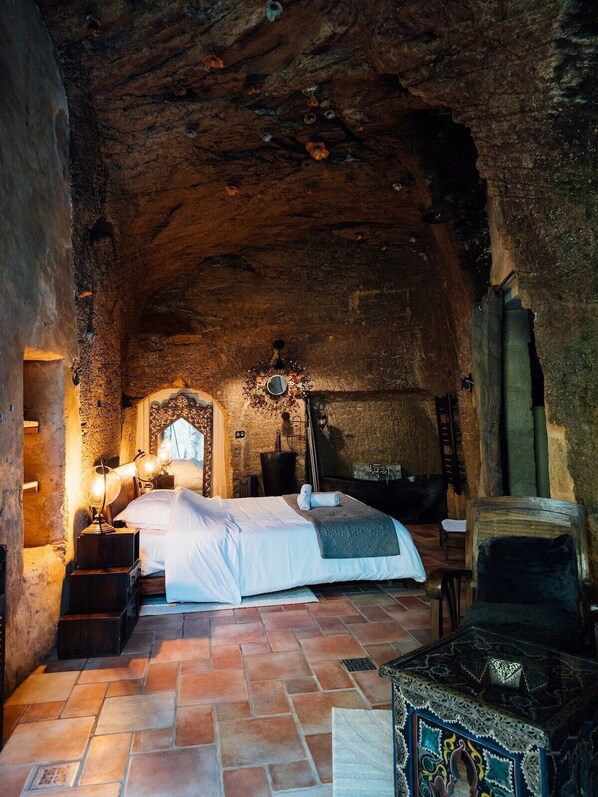 A rustic, stone-walled room with a large bed, a wooden chest, and a stone bench.
