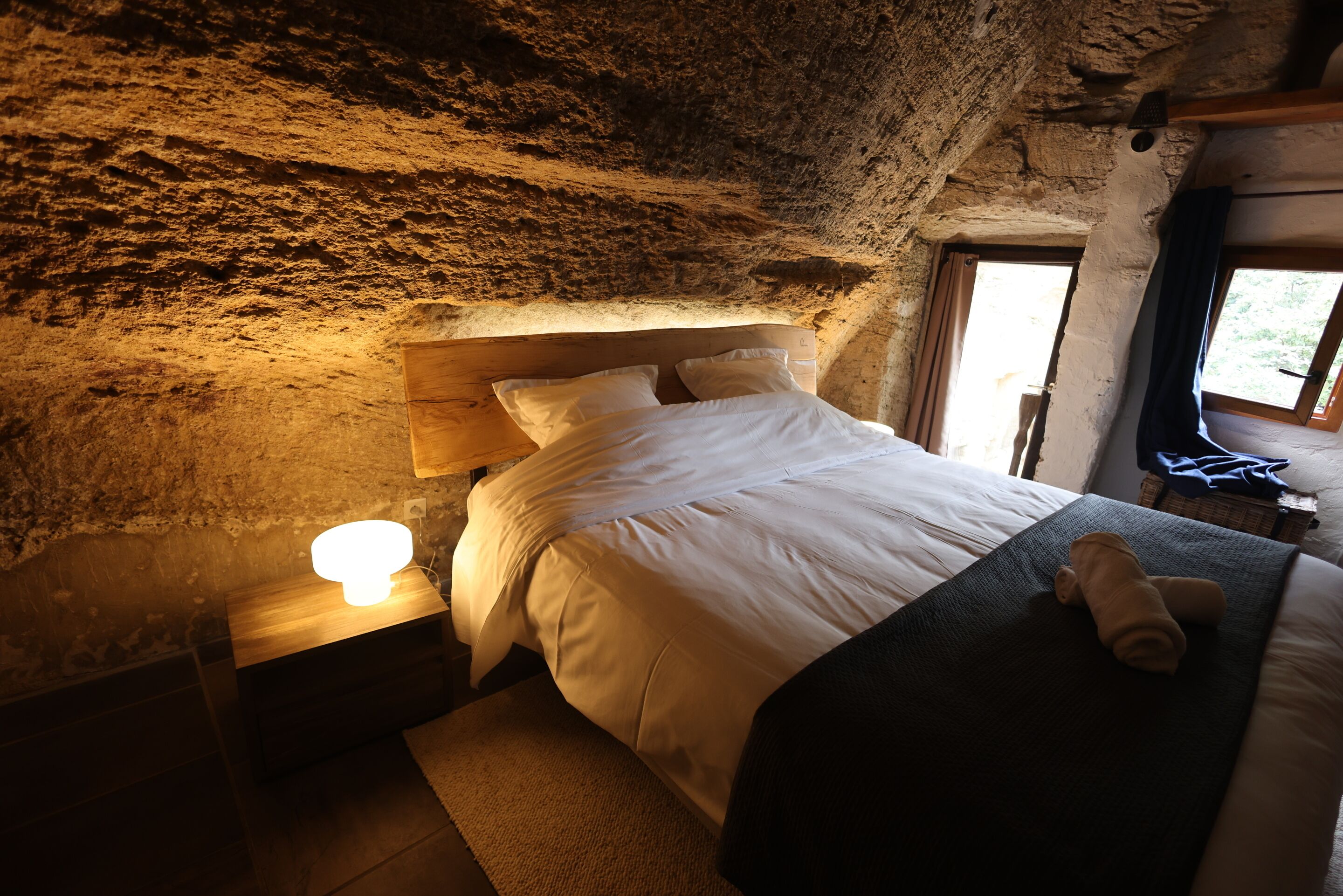 A cozy bedroom with a rustic stone ceiling, a wooden headboard, a bedside lamp, and a neatly made bed with a rolled towel.