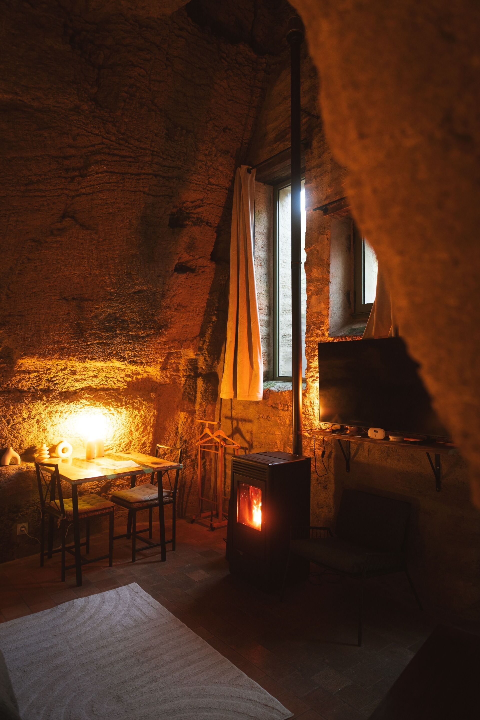A cozy room with a stone wall, a wooden table, a chair, a window with curtains, and a wood-burning stove.