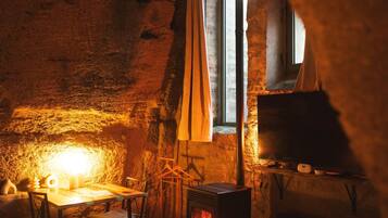 A cozy room with a stone wall, a wooden table, a chair, a window with curtains, and a wood-burning stove.