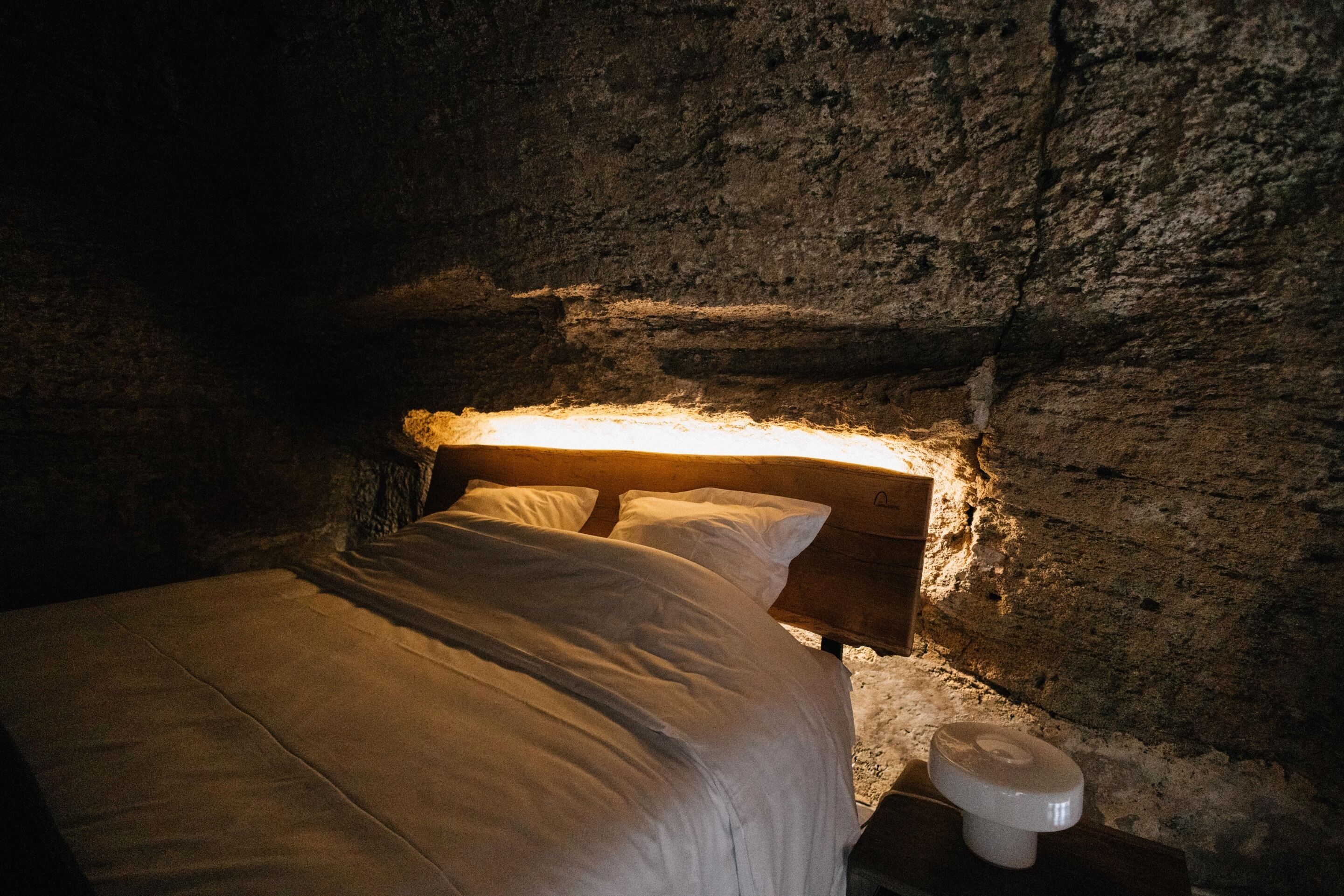 Un lit dans une pièce aux murs de pierre, avec une tête de lit en bois et un petit objet blanc sur le sol.