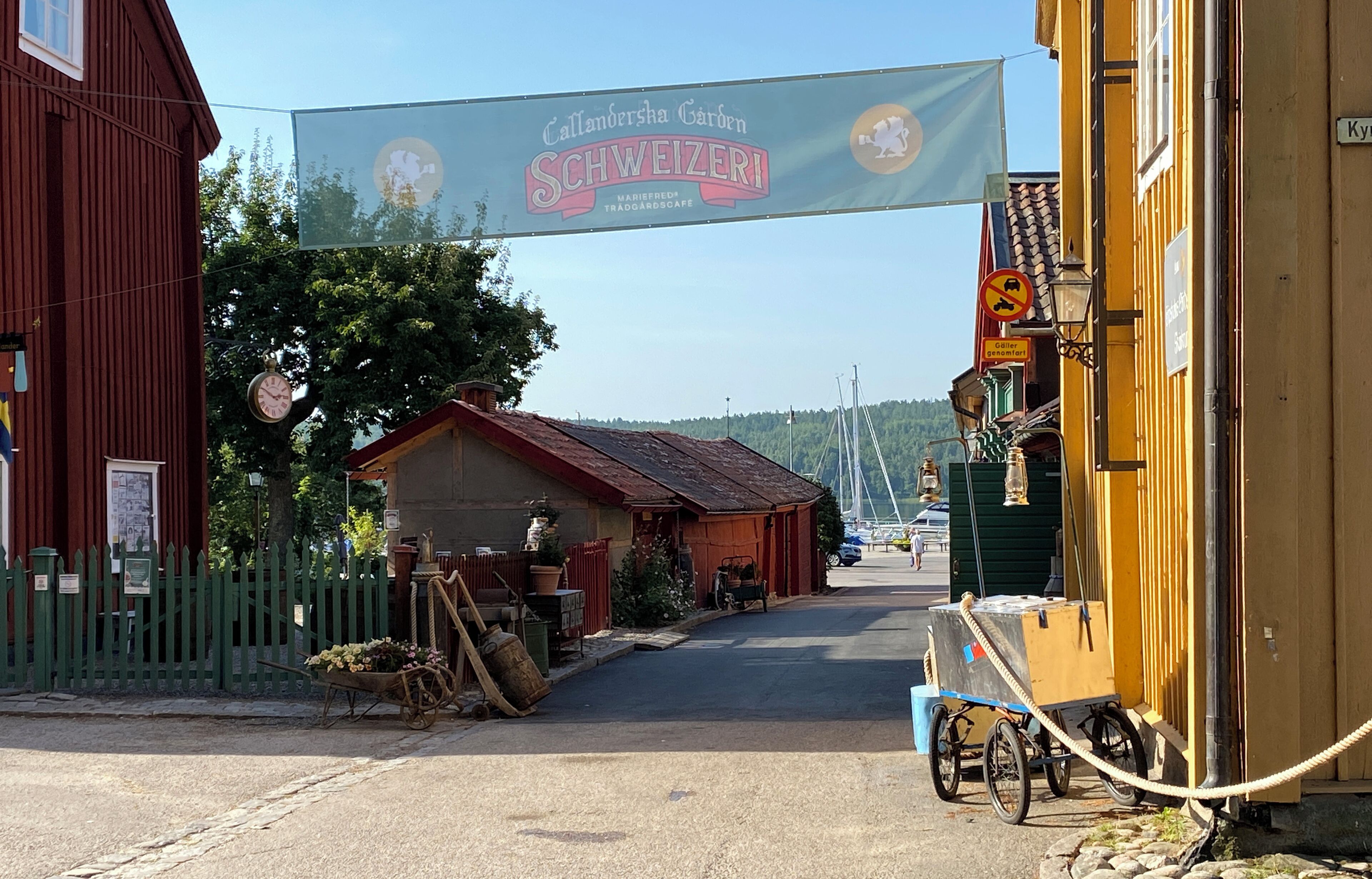 Vista de la calle con un cartel anunciando 'Callanderscha Garden' y el restaurante 'Schweizeri', una cerca verde, un carretón y un edificio amarillo.