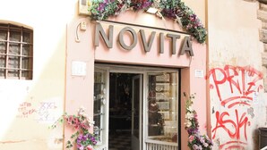En butik med navnet NOVITA, der har et skakmønstret gulv, en træbænk udenfor og blomsterudsmykning.