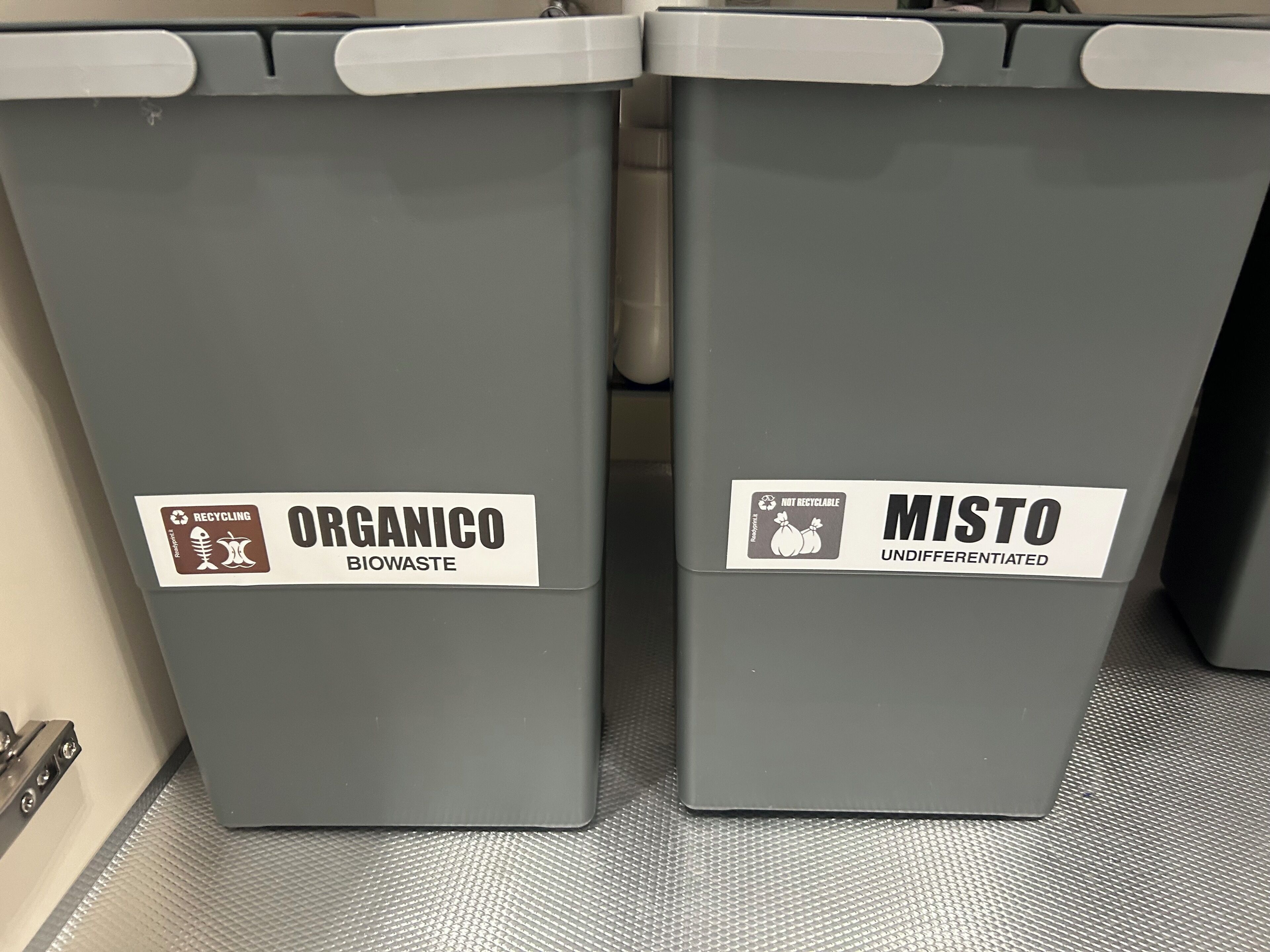 Two gray waste bins labeled 'Organico Biowaste' and 'Misto Undifferentiated' with recycling symbols.