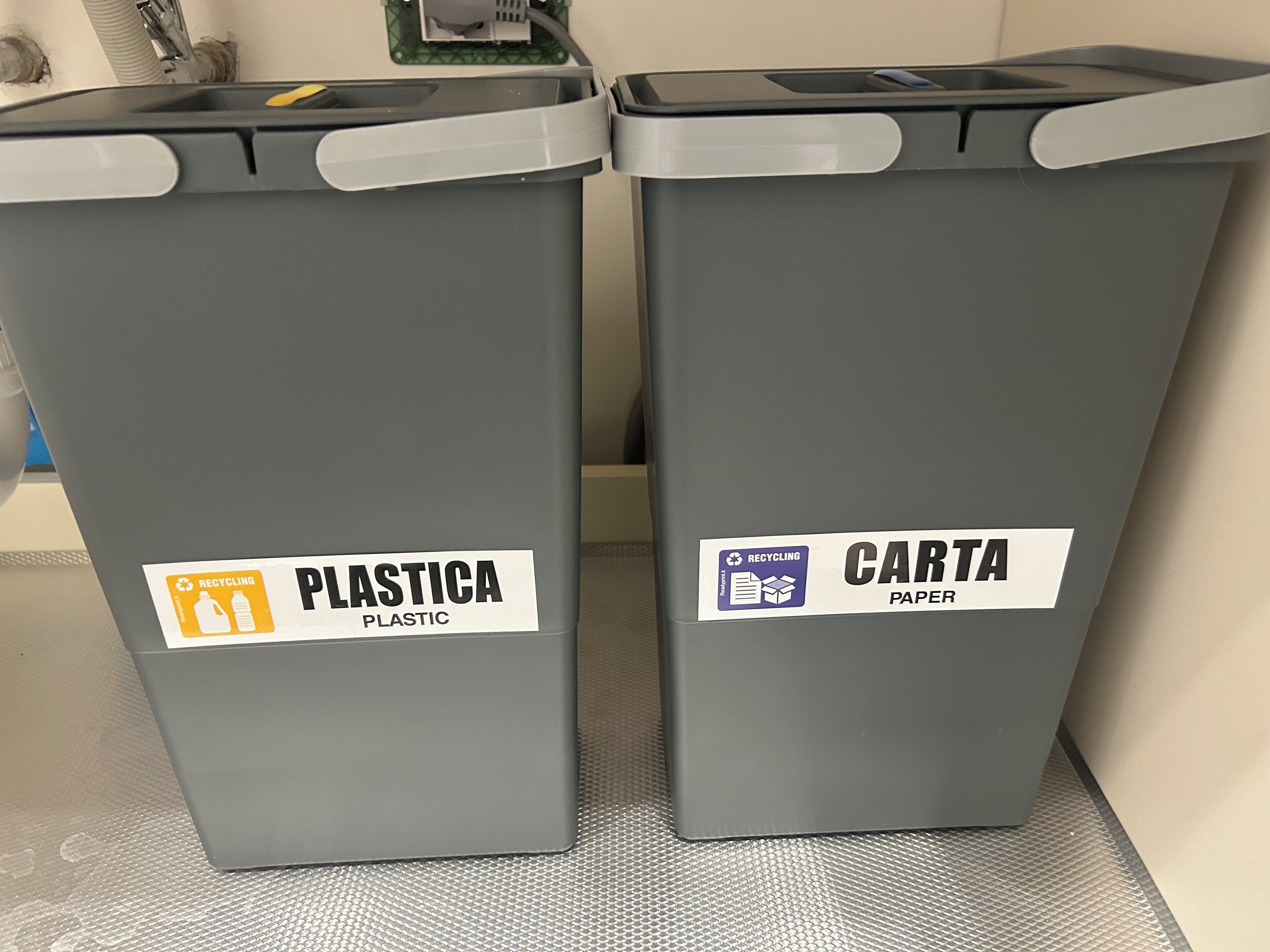 Zwei graue Recyclingbehälter, einer beschriftet mit 'PLASTICA' für Kunststoff und der andere mit 'CARTA' für Papier, stehen auf einem strukturierten Boden.