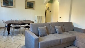 A living room with a grey sofa, a dining table with clear chairs, and two framed paintings on the wall. - Casa dei fiori appartamento  6 persone - by Ghor (Rome)
