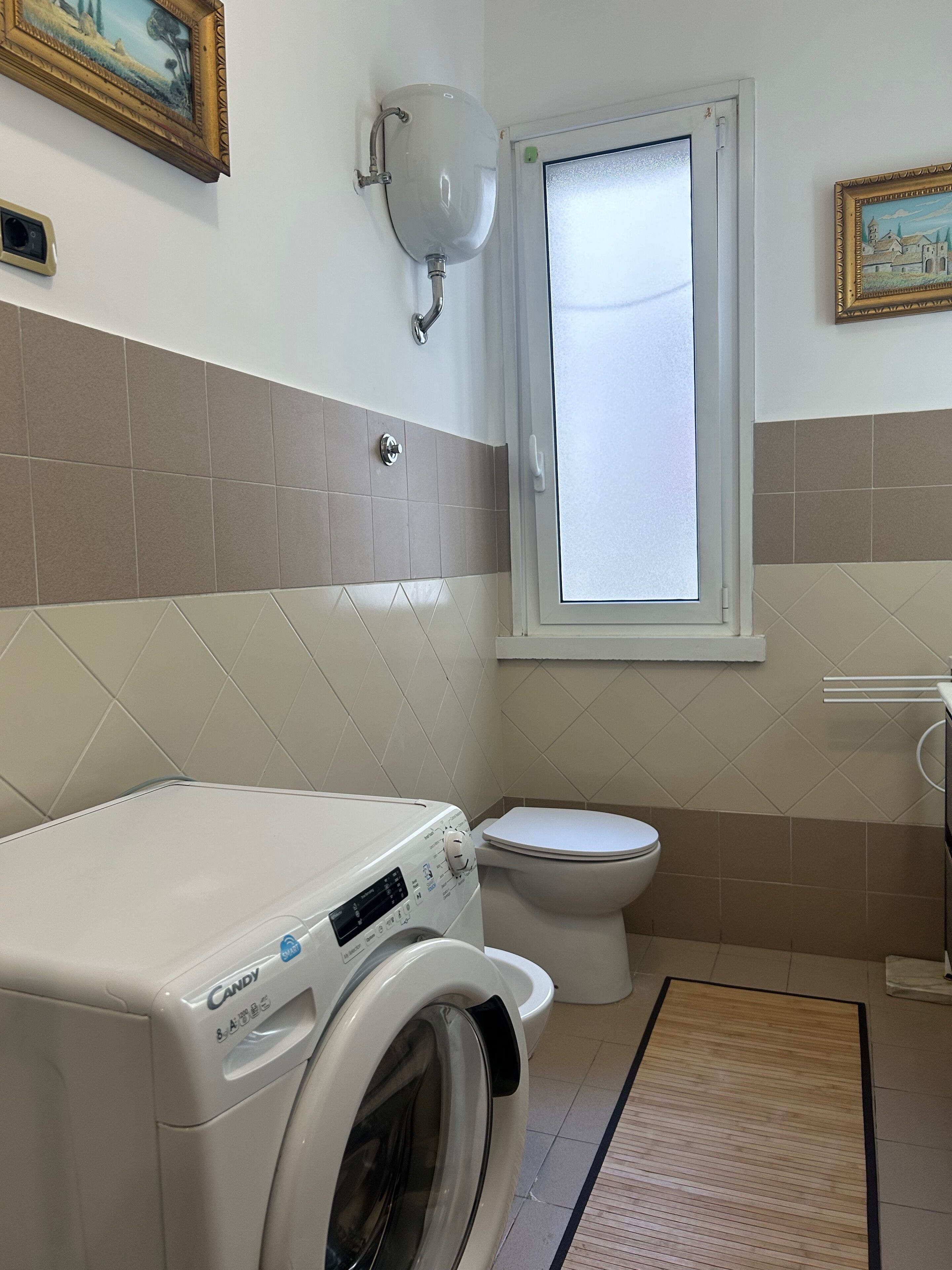 Een kleine badkamer met een wasmachine, toilet en een raam met binnengeborsteld glas.