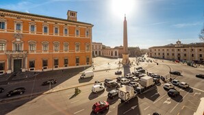 Ein belebter Stadtplatz mit einem markanten Obelisken, umgeben von historischen Gebäuden und blauem Himmel.